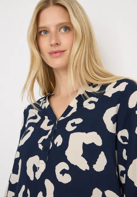 Frau mit blonden Haaren trägt eine dunkelblaue Bluse mit beigem Muster.